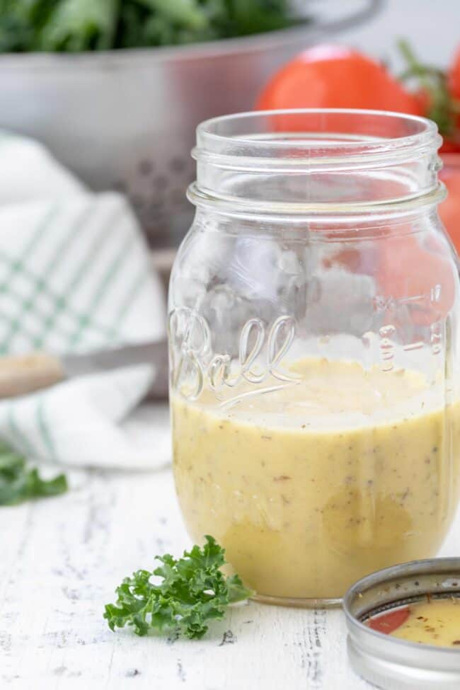 A clear mason jar with homemade a vinaigrette. A metal colander filled with lettuce and tomatoes in the background.