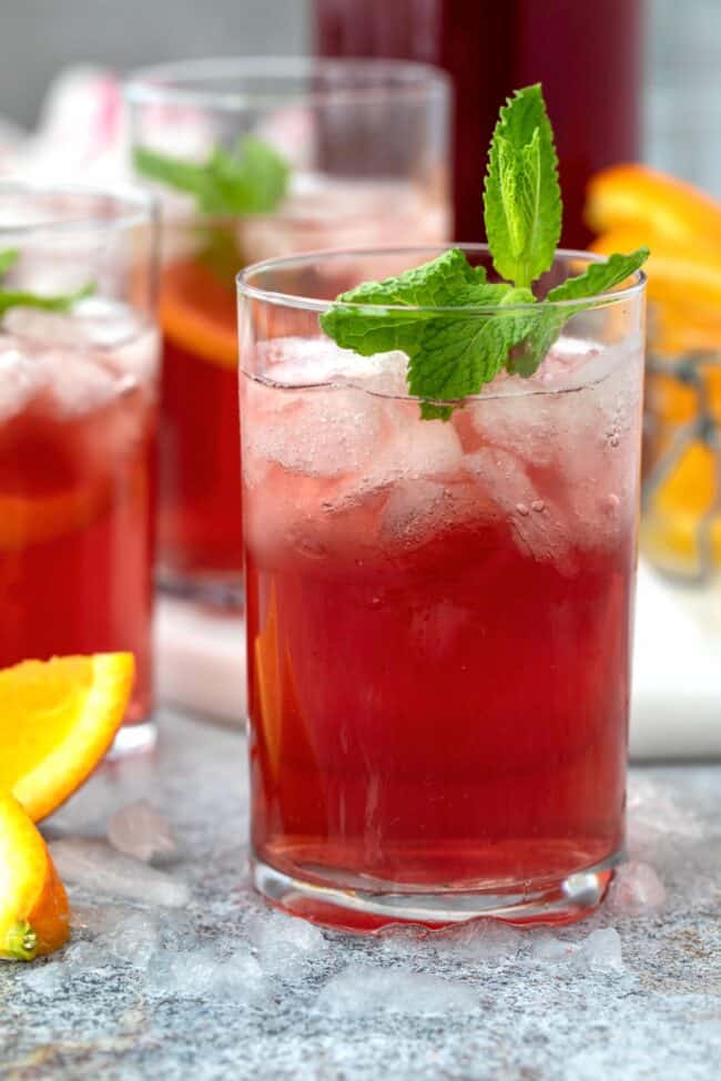 Clear drinking glasses filled with apple cider vinegar and cranberry juice and ice. Wedges of orange sit next to the glasses.