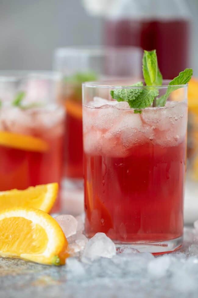 Three clear drinking glasses filled with apple cider vinegar and cranberry juice and crushed ice. Orange wedges and crushed ice are scattered next to the glasses.