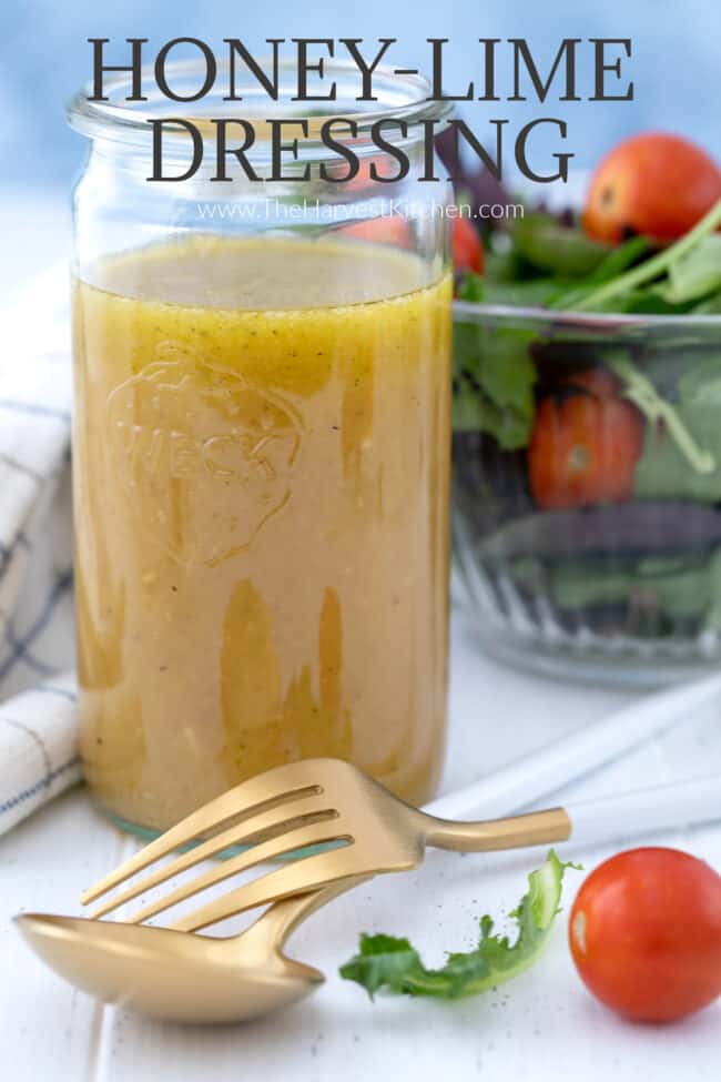 Glass mason jar filled with honey lime dressing. Glass bowl filled with salad in the background with fork and spoon.