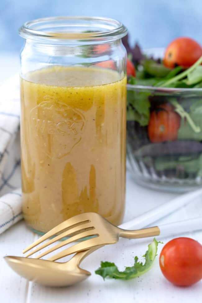 Glass mason jar filled with honey lime dressing. Glass bowl filled with salad in the background with fork and spoon.