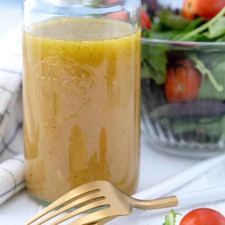Glass mason jar filled with honey lime dressing. Glass bowl filled with salad in the background with fork and spoon.
