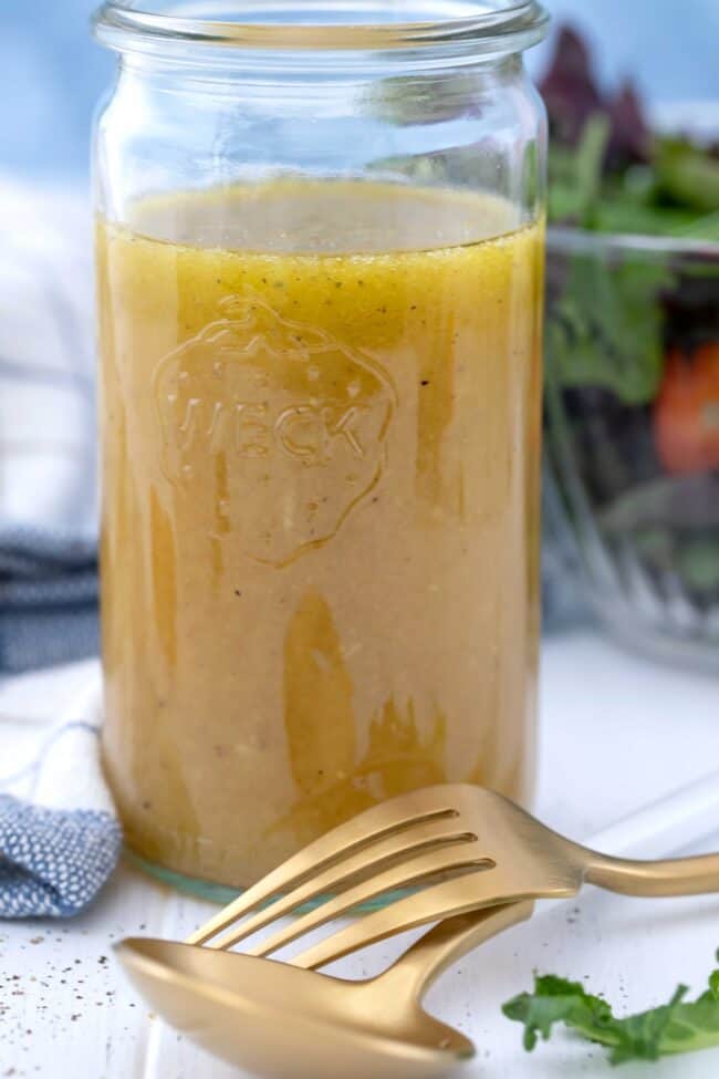 A clear glass mason jar filled with honey live dressing. A salad is in a glass bowl behind the mason jar, and a fork and spoon sit next to the jar.