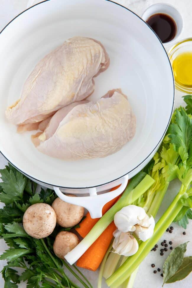 Boneless chicken breasts sit in a white pot. Celery, carrots, mushrooms and garlic sit next to the pot.