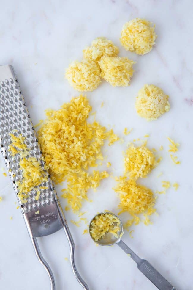 A metal zester sits next to a pile of lemon zest on a white cutting board,