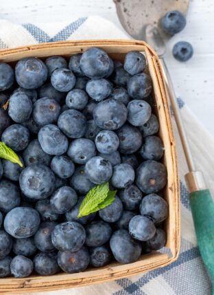 A wooden fruit basket filled with blueberries. A spoon sits next to the basket.