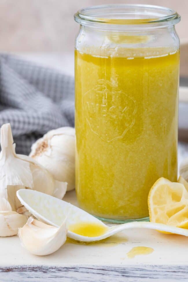 A clear glass jar of garlic oil. A white spoon rests next to the jar.