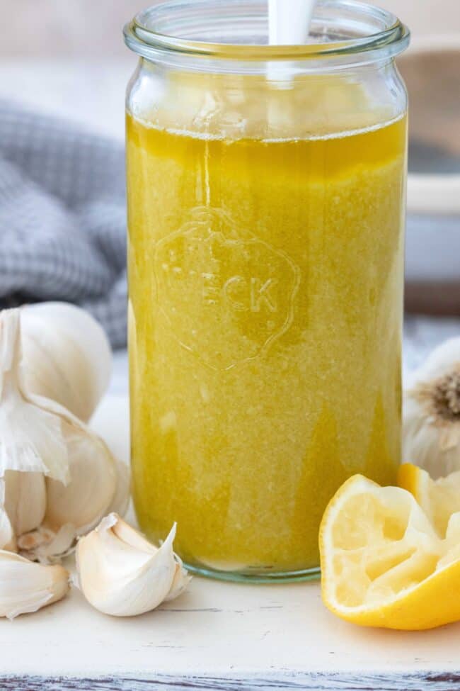 A clear glass jar of Lemon Garlic Oil. Garlic cloves and wedges of lemon set next to the jar. 