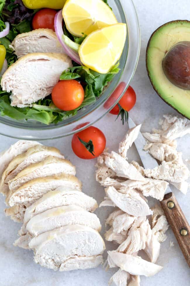 Slices of poached chicken and shredded pieces of shredded chicken sit on a cutting board next to a knife.