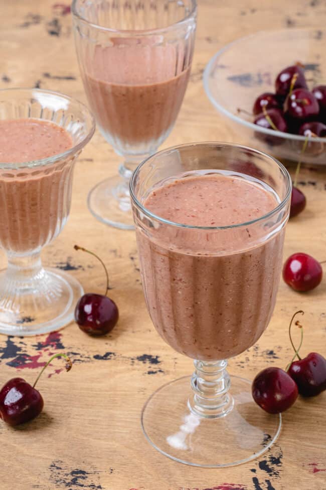 3 glasses filled with chocolate cherry oat smoothie. Red cherries are scattered around the glasses.