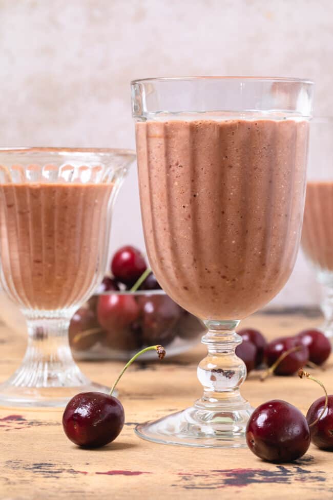 3 glasses filled with chocolate cherry oat smoothie. Red cherries are scattered around the glasses.