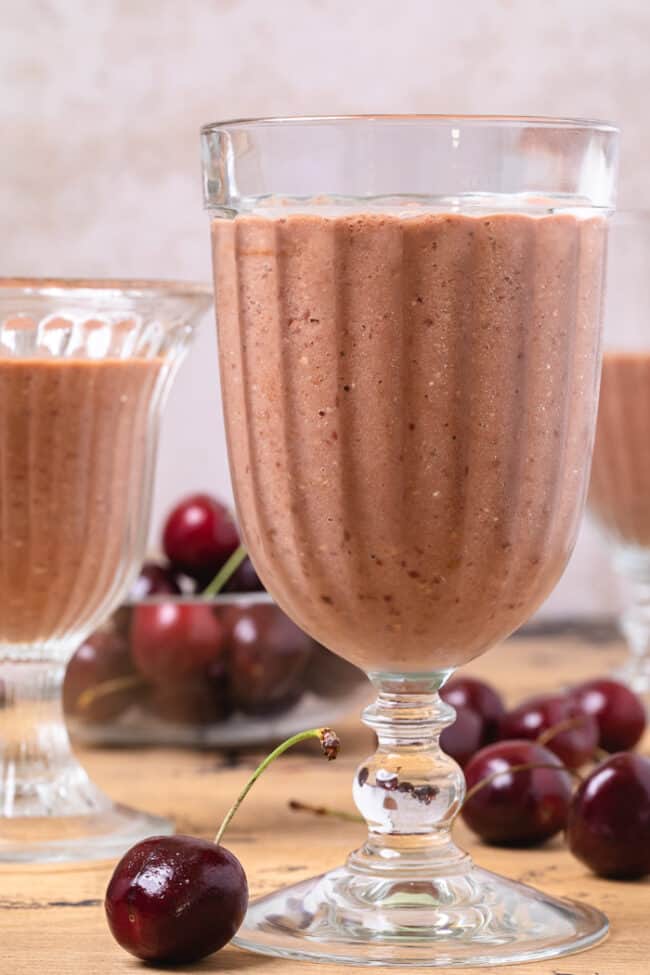 3 glasses filled with chocolate cherry oat smoothie. Red cherries are scattered around the glasses.