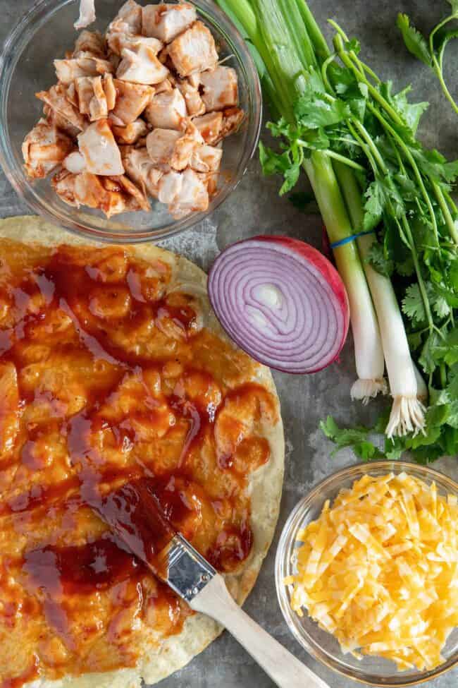 A flour tortilla with bbq sauce on it, a clear glass bowl filled with chopped cooked chicken and one filled with grated cheese.