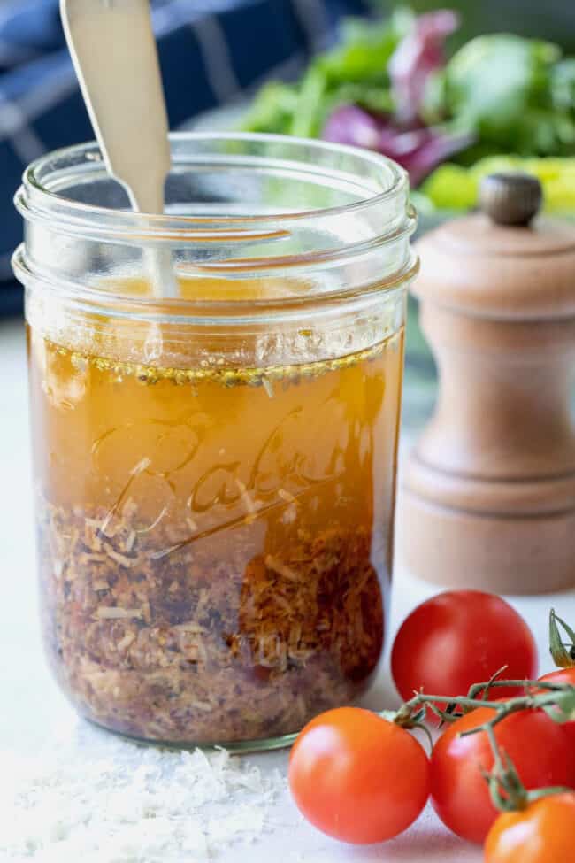 A clear glass mason jar filled with oil, parmesan cheese and seasoning. Cherry tomatoes lay next to the jar with a salad in the background.