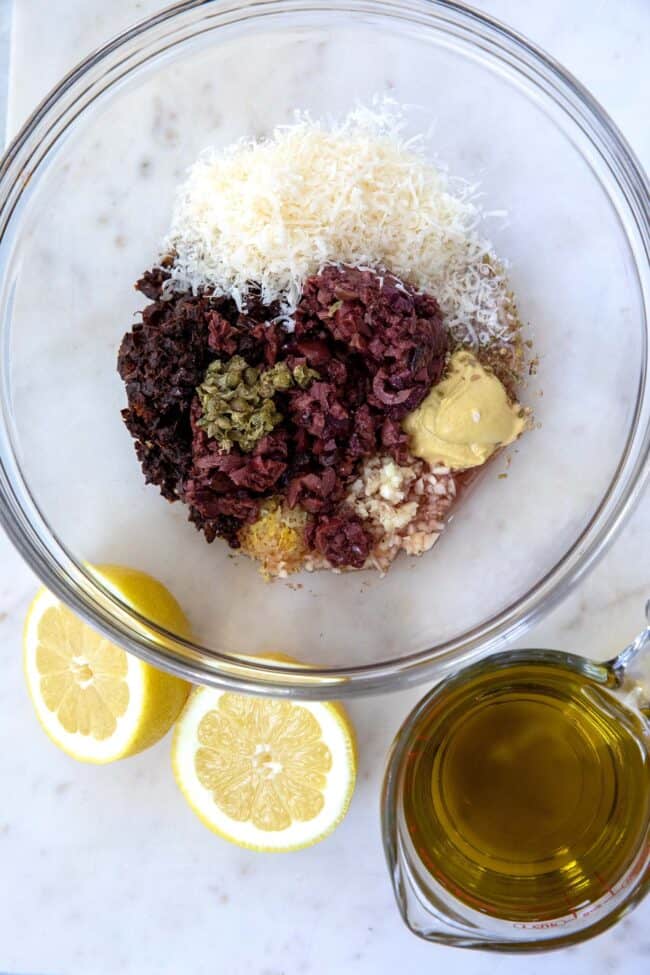 A clear glass mixing bowl filled with kalamata olives, mustard, red wine vinegar and parmesan cheese. Lemon halves are next to the bowl with a glass measuring cup filled with olive oil.