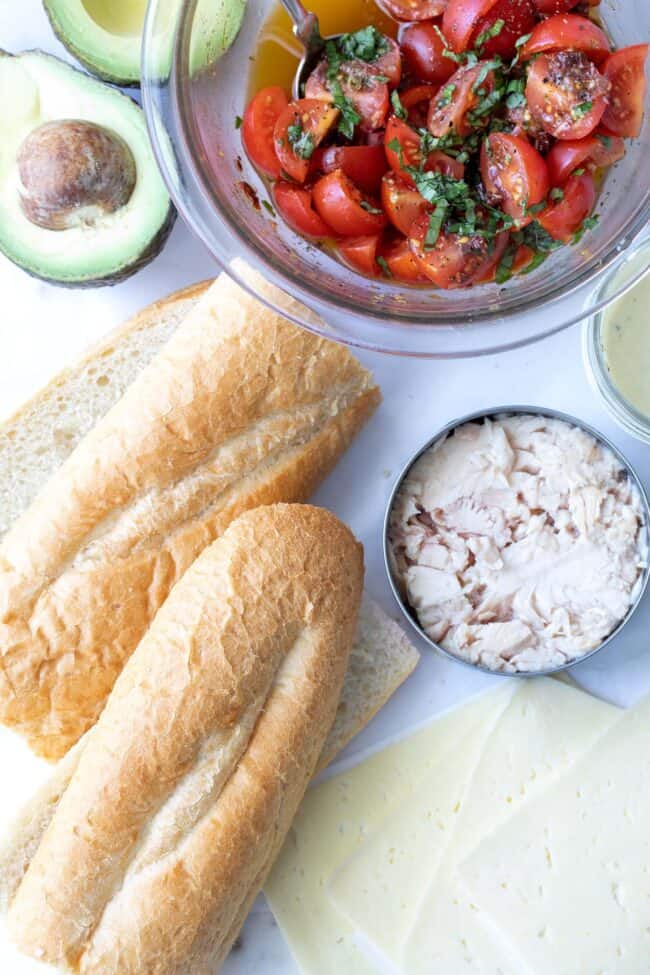 A baguette, cheese slices and a bowl of marinated cherry tomatoes.