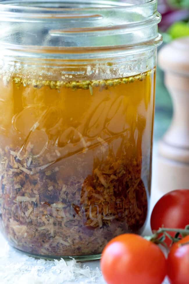 A clear glass mason jar filled with Greek salad dressing. Cherry tomatoes lay next to the jar.