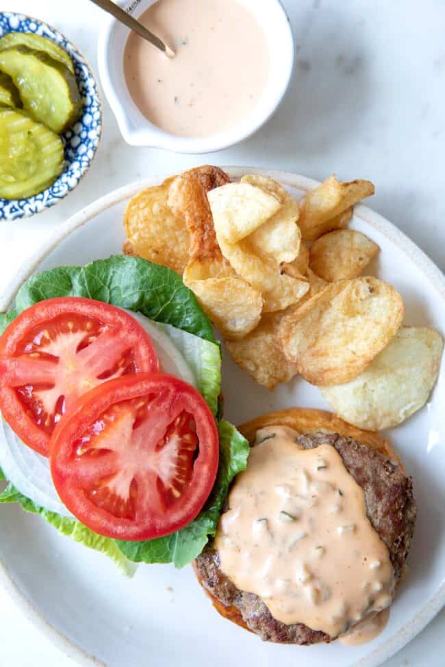 A white plate with burger on a bun with tomatoes, lettuce and potato chips.