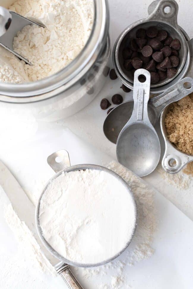a glass jar filled with all-purpose flour sits next to silver measuring cups filled with chocolate chips, brown sugar and flour