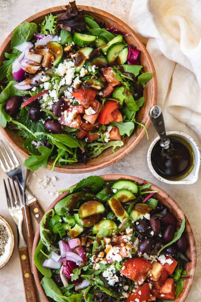 two wooden bowls filled with Mediterranean salad. Three forks are to the left of the bowls and a bowl with balsamic vinaigrette sits to the right of the bowls.