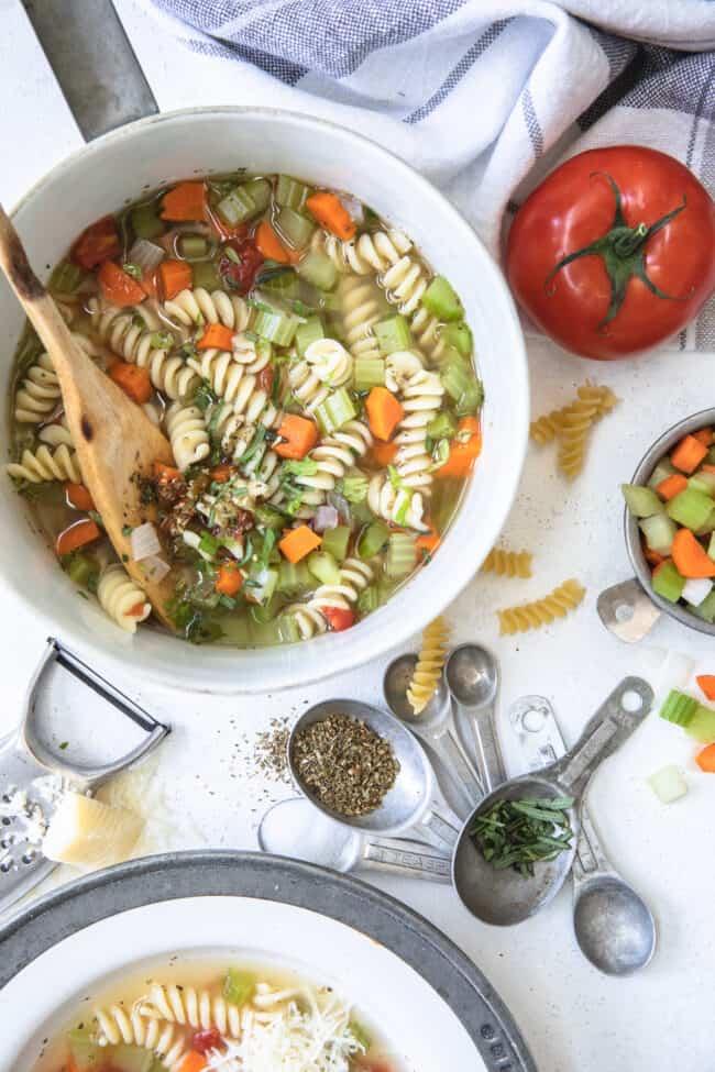 to represent how many teaspoons in a tablespoon (tsp to tbsp) there is a white pot filled with vegetable noodle soup. A mixture of measuring spoons filled with different herbs sits next to the po.