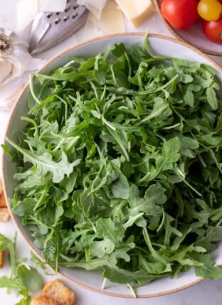 A white bowl of arugula with cherry tomatoes and parmesan cheese next to it