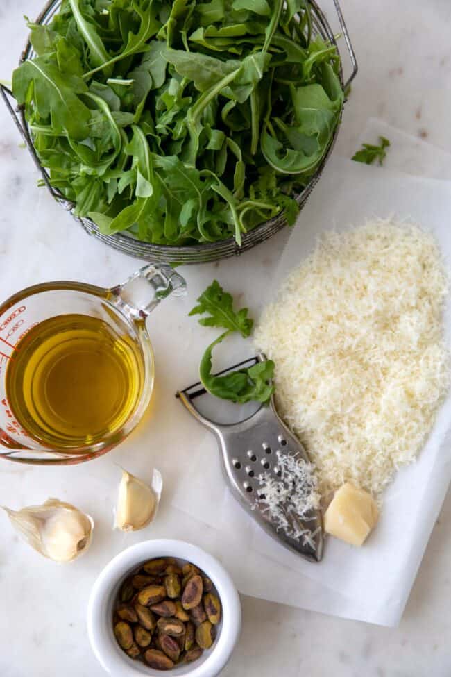 A basket of fresh arugula with grated Parmesan cheese next to it. A measuring cup with olive oil, 2 garlic cloves and a small dish filled with pistachio nuts.