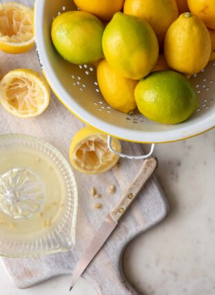 A white colander filled with lemons. A glass juicer filled with lemon juice sits next to the colander.