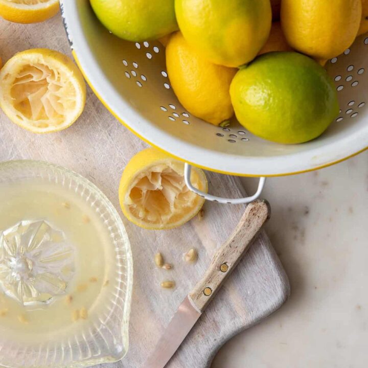 A white colander filled with lemons. A glass juicer filled with lemon juice sits next to the colander.