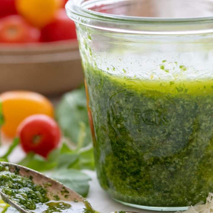 A glass jar filled with Arugula Pesto. A spoon with pesto in it sits next to the jar.