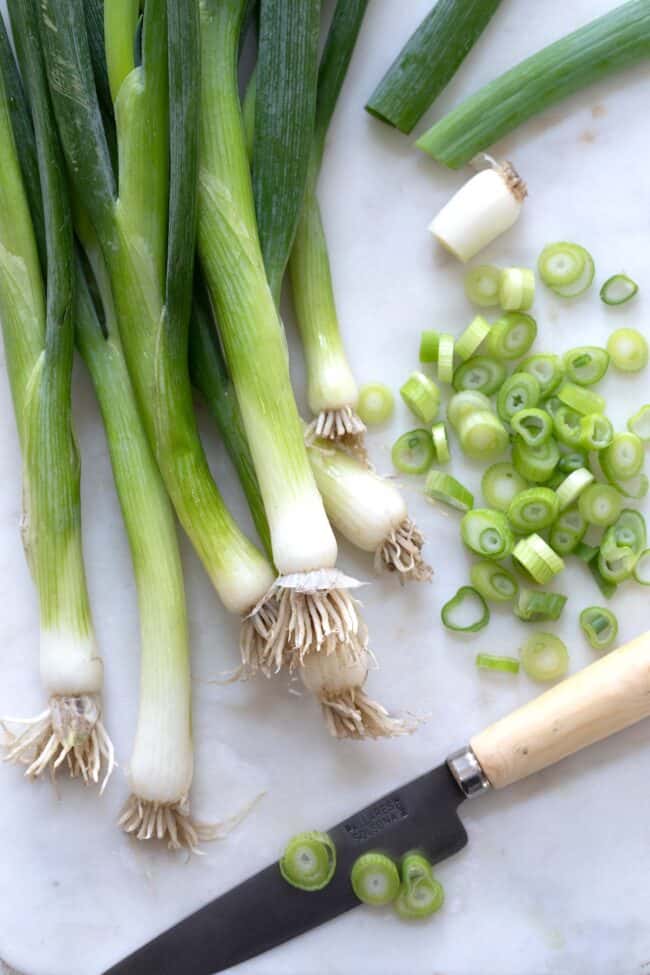 A bunch of green onions are on a cutting board. A kitchen knife sits next to them and a small pile of chopped green onions.