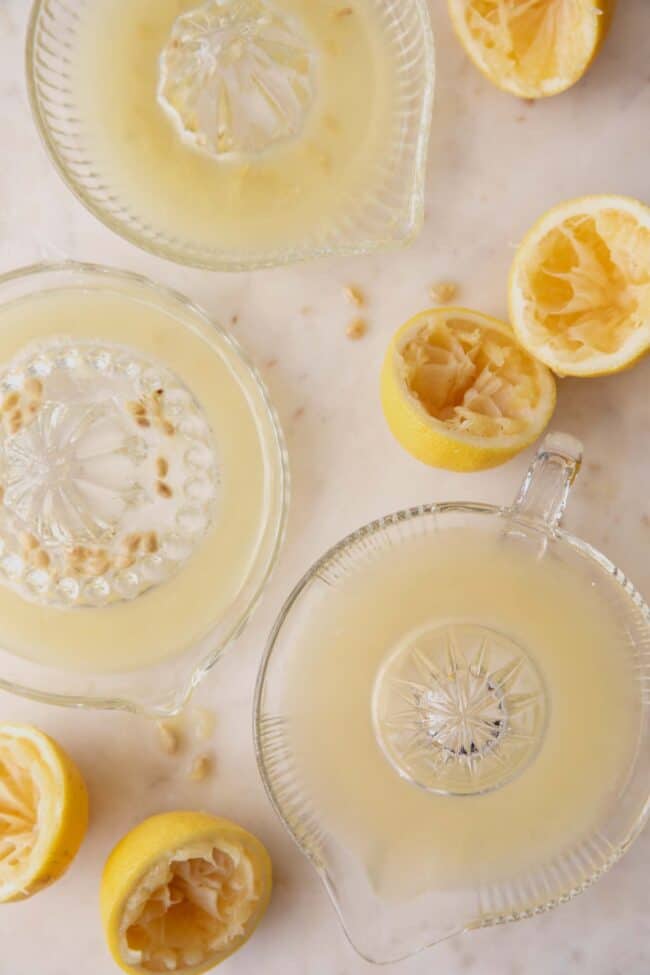 3 glass juice presses filled with lemon juice. Lemons cut in half and juiced sit next to them.