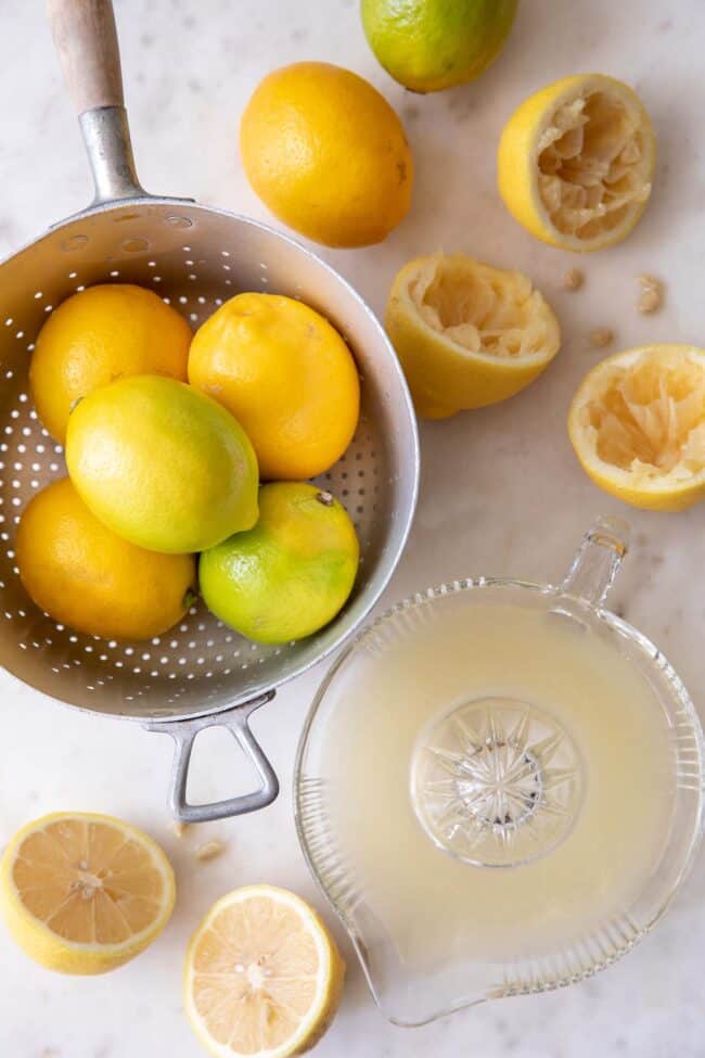 A silver colander filled with lemons. lemons cut in half and juiced in a glass juicer sit next to the colander. For tips and tricks on juicing lemons and how much juice in one lemon