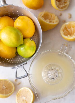 A silver colander filled with lemons. lemons cut in half and juiced in a glass juicer sit next to the colander. For tips and tricks on juicing lemons and how much juice in one lemon