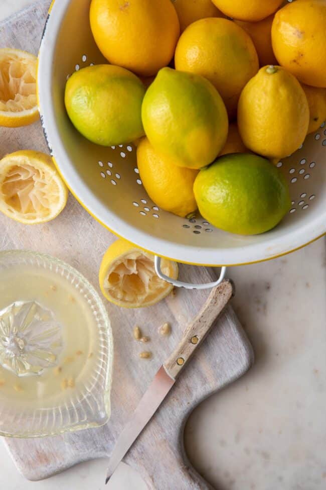 A white colander filled with lemons. A glass juicer filled with lemon juice sits next to the colander.