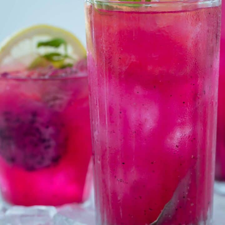 two glasses filled with mango dragonfruit green tea drink. Mint leaves and sliced lemon poking out of drinks.