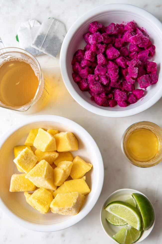 SEVERAL white bowls filled with frozen mango and dragonfruit and cut limes. A glass filled with honey and a glass filled with green tea next to the bowls of fruit.