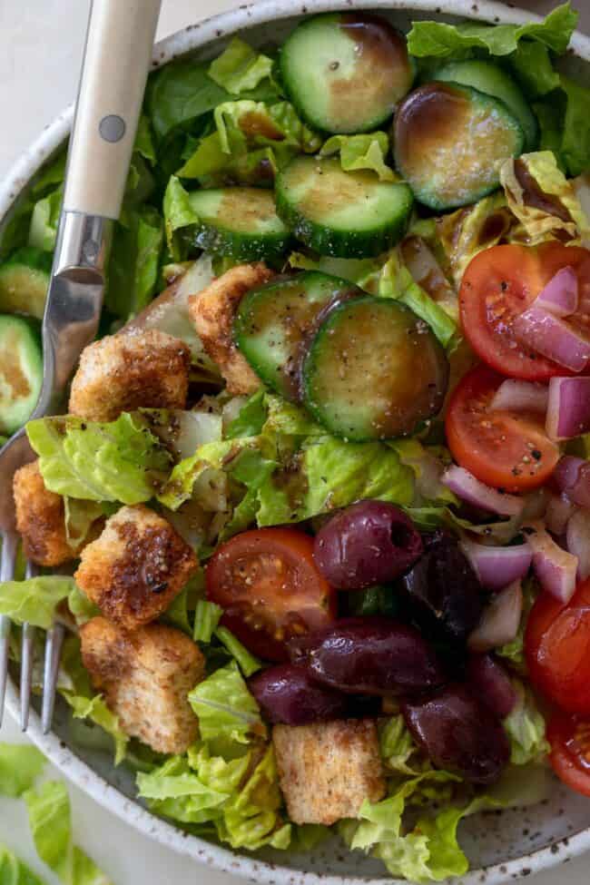 A white bowl of leafy greens with cucumbers, tomatoes, red onion, kalamata olives and croutons drizzled with balsamic vinaigrette. 