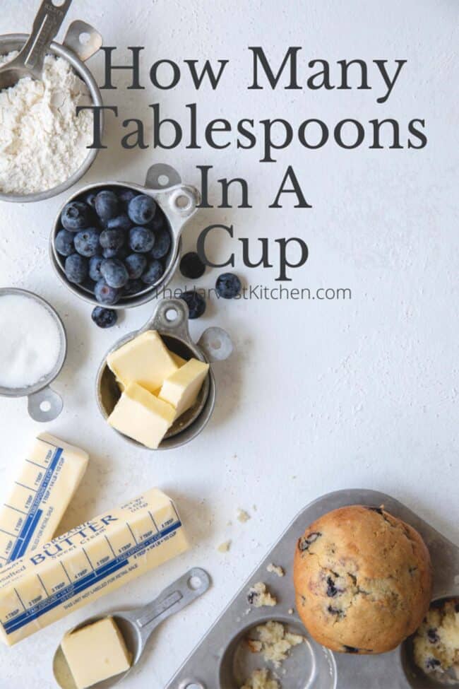 Vintage aluminum measuring cups filled with flour, butter, sugar and blueberries to learn how many tablespoons in a cup (tbsp to oz). A muffin tin sits next to them with a baked blueberry muffin.