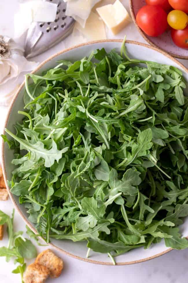 A white bowl of arugula with cherry tomatoes and parmesan cheese next to it