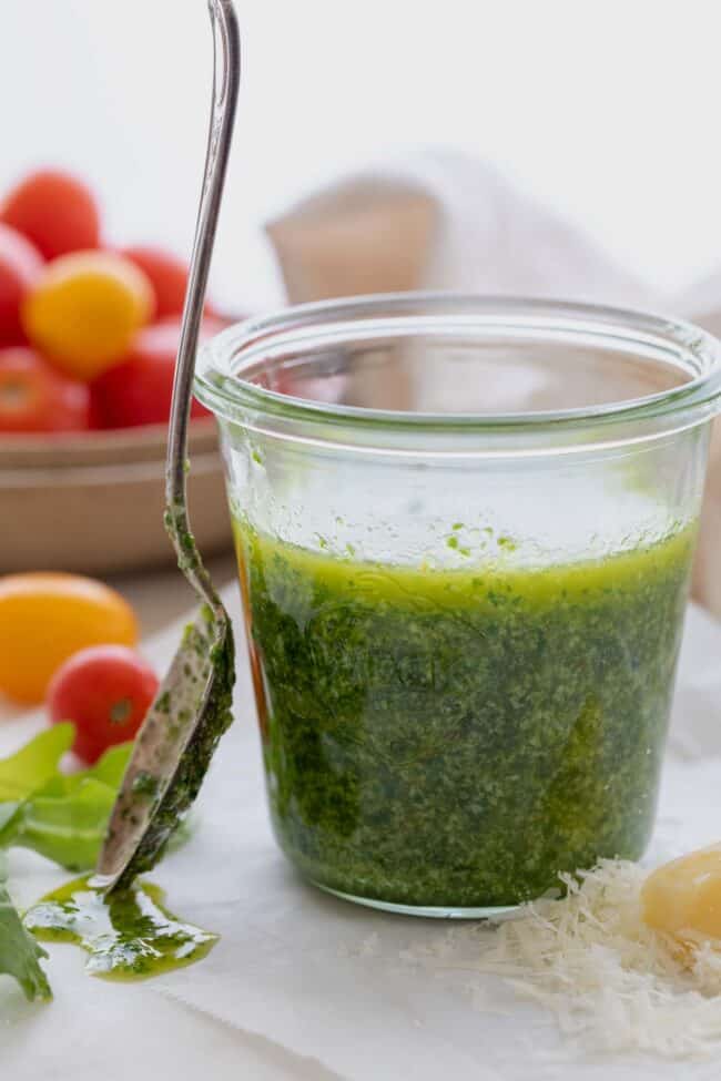 A glass jar filled with arugula pesto (pistachio pesto). A spoon with pesto in it sits next to the jar.