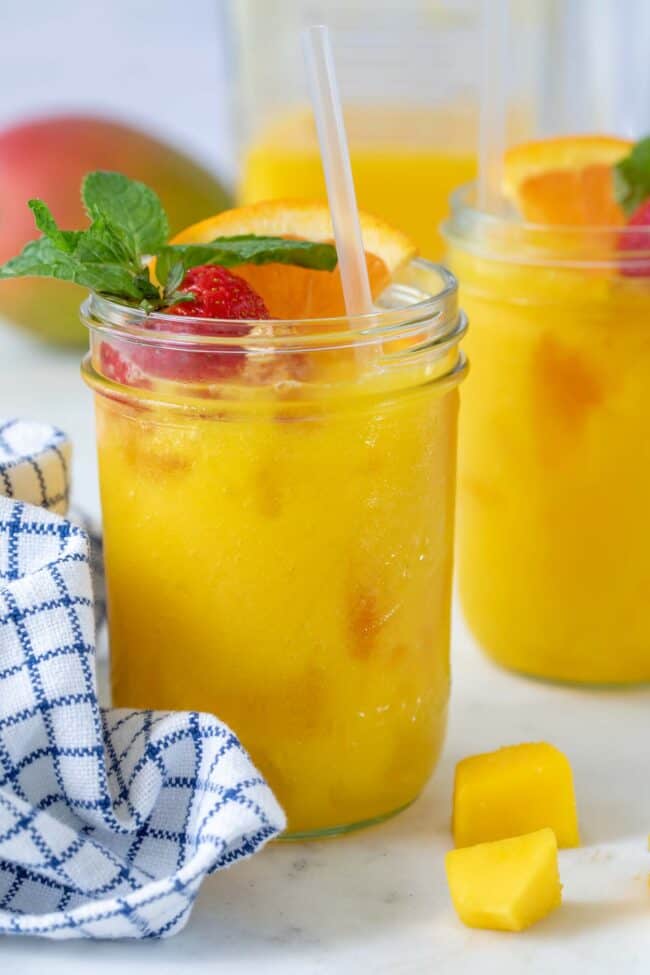 Two glasses filled with orange juice and a straw. Slices of range and strawberry are placed on top of the ice in the glasses. A blue and white dish towel sit next to the glasses.