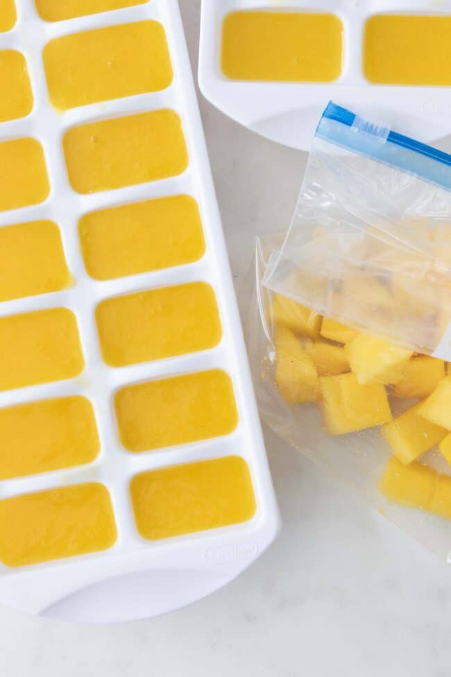 A white ice cube tray filled with mango nectar (king of fruit) ice cubes. A plastic bag filled with cut pieces of mango sits next to the white tray.