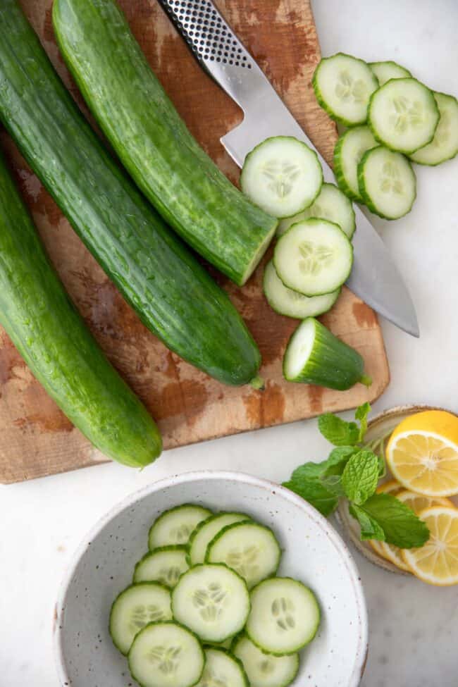 Three English cucumbers on a wood cutting board with a knife. A bowl of sliced cucumbers sits next to the cutting board.