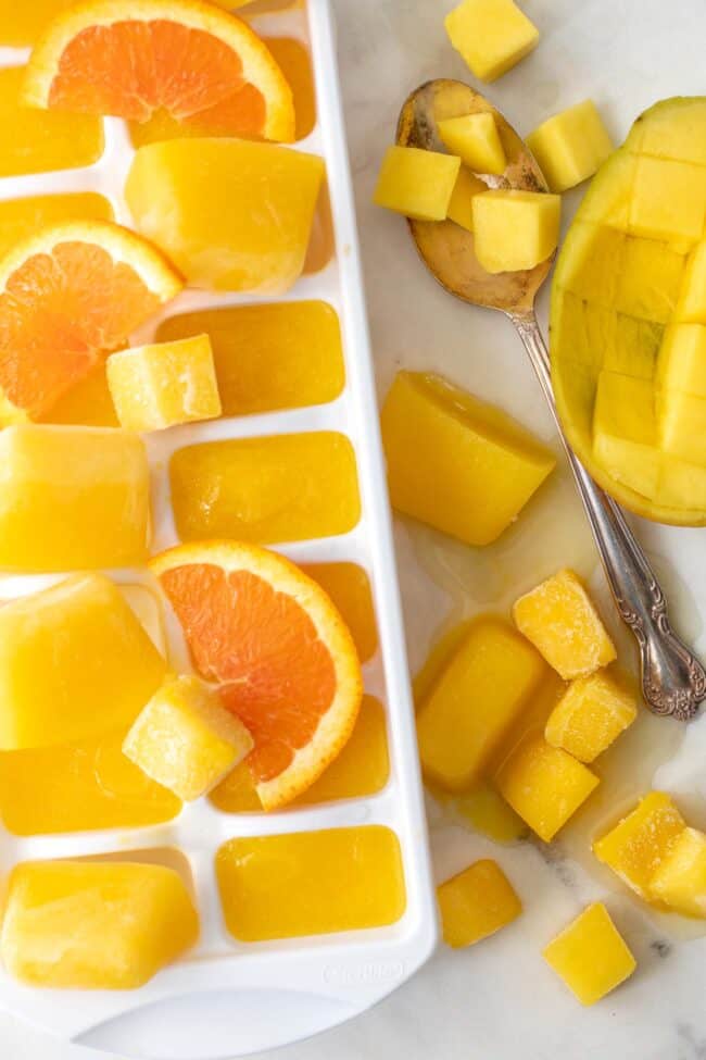 white ice cube tray filled with mango nectar juice ice cubes. Cut mango pieces sits next to the tray.