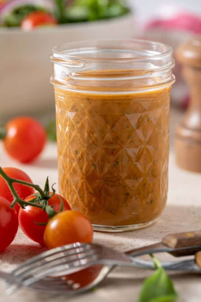 Mason jar filled with sun dried tomato vinaigrette. A bowl with salad and two forks sit next to the mason jar.