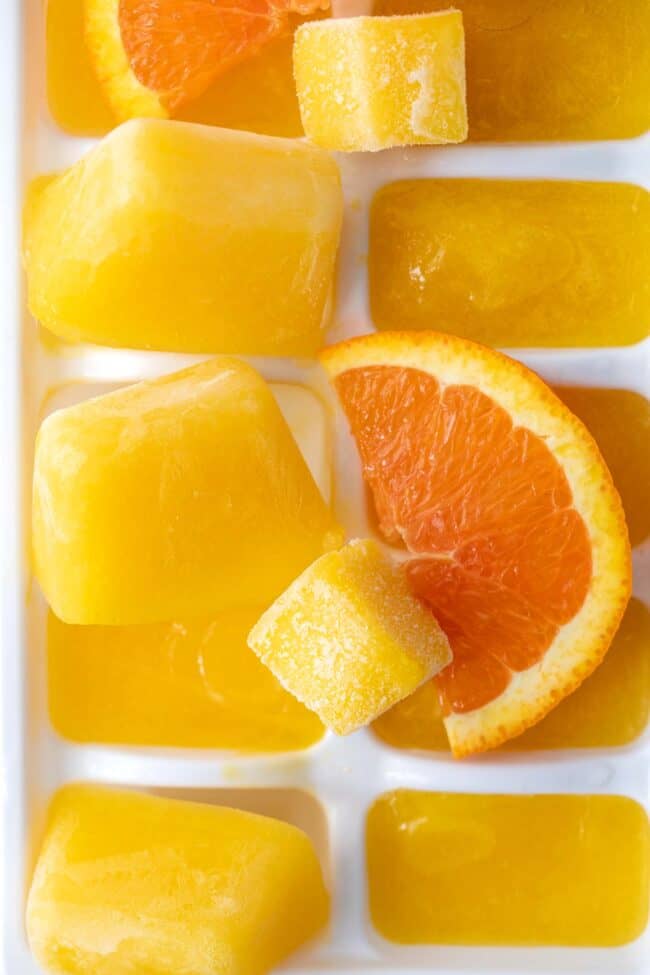 A white ice cube tray filled with orange ice cubes. Two slices of oranges sit on top of the tray.