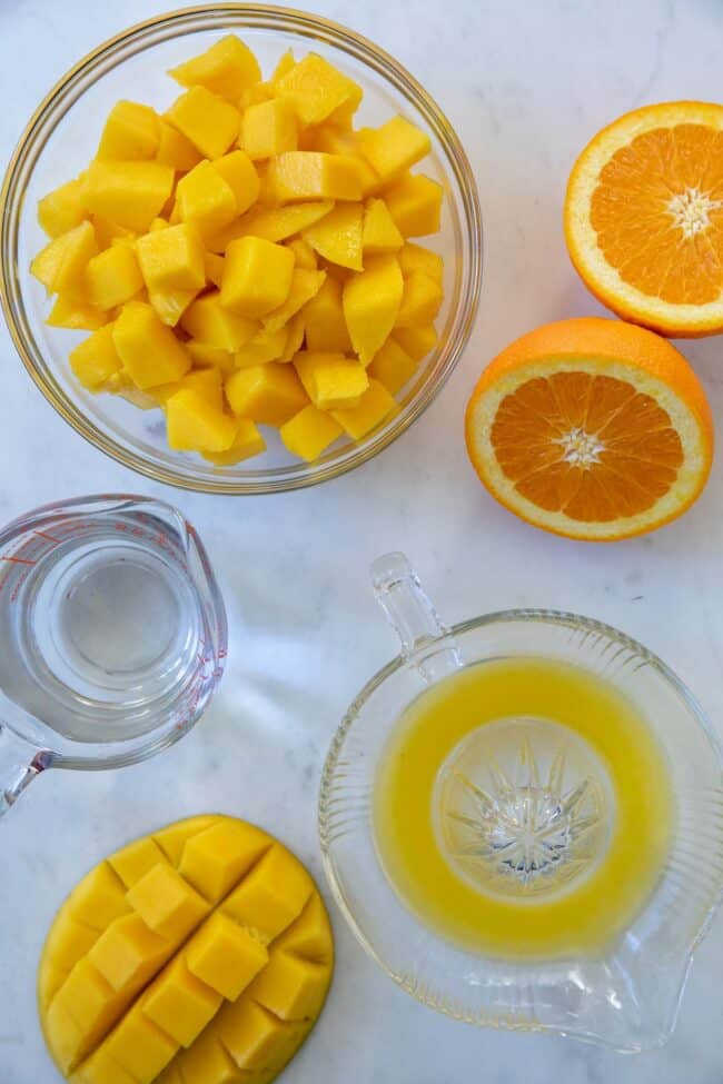 A glass bowl filled with cut mango, with an orange cut in half sitting next to is. Half of a mango cut sits next to a juicer filled with some orange juice. 