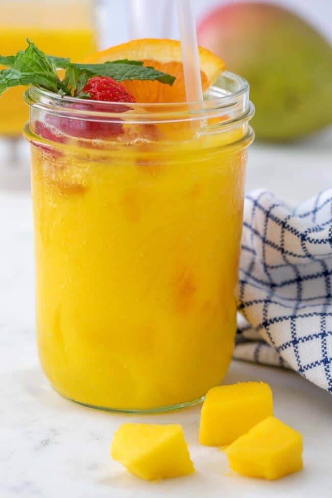 A drinking glass filled with mango juice with a sprig of mint, slice of orange and a strawberry are garnished on top. There is a blue and white towel next to the glass.