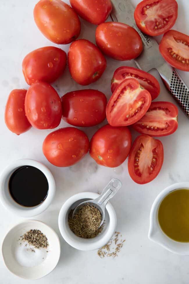 Roma tomatoes set on a white counter with a knife. White ramekins are filled with olive oil, balsamic vinegar and seasonings.
